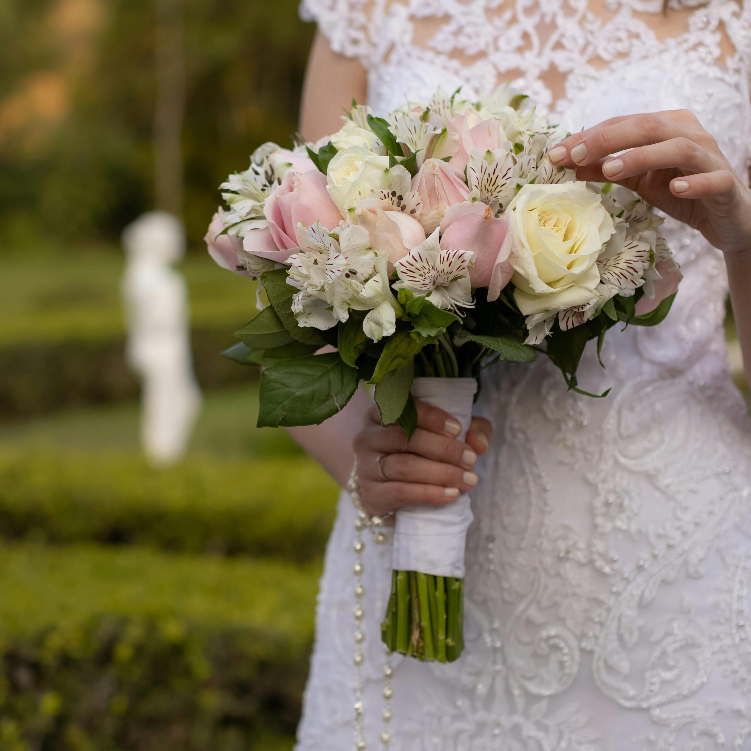 A bride holding an elegant bouquet in a lace dress outdoors in a garden setting.